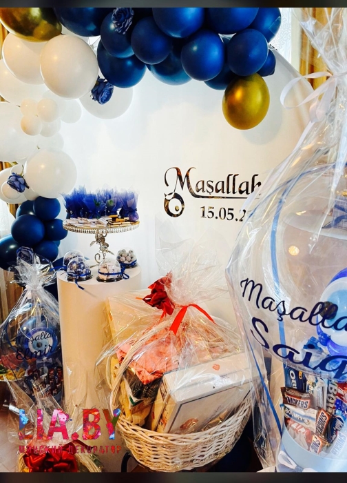 Фотозона в виде белой круглой ткани с разноразмерной гирляндой из синих и белых шаров, перед которой находятся декорации в виде подарков и еды