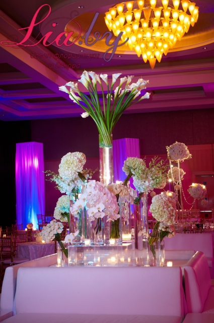 Калы в высокой вазе, композиция оформлена цветочными вставками у подножья конструкции. Множество цилиндров, цветов и стеклянное основание цветочной инсталляции.