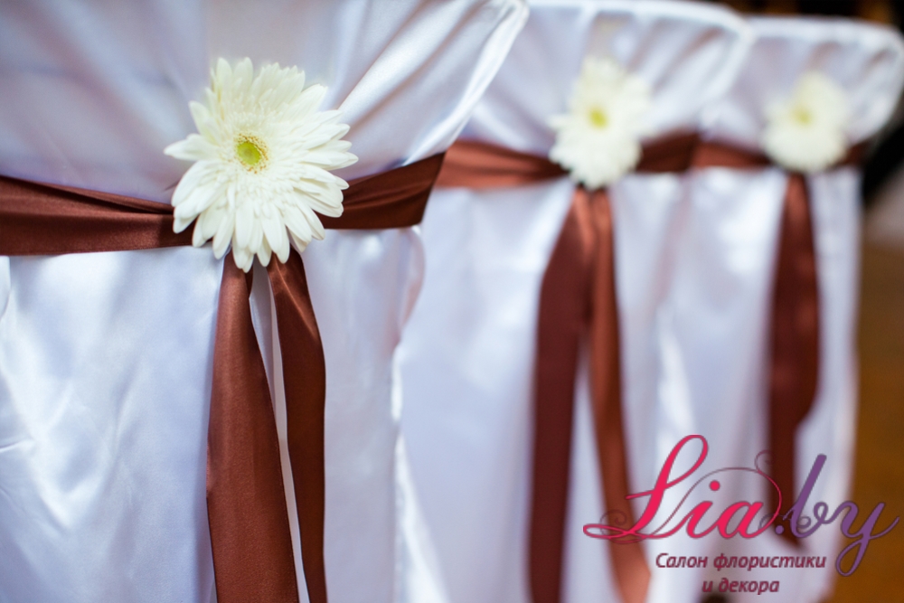 Стулья украшены полным белым чехлом и обвязаны атласной коричневой лентой с цветком-брошью