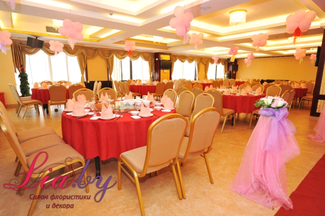 Свадебный банкет, на котором столы и стулья не украшены, под потолком зала гелиевые шары. Стойка ведущего украшена фатином и цветами