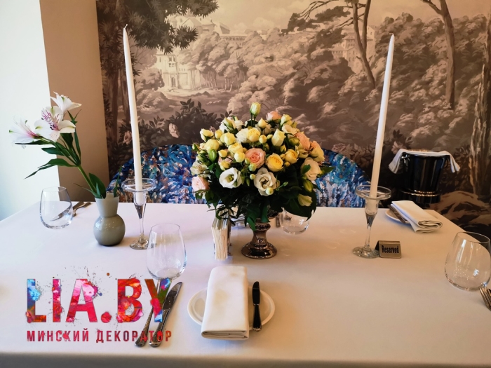 украшение столика в ресторане пышной цветочной композицией из натуральной розы, эустомы и зелени в белых и нежно желтых тонах, подсвечниками с высокими белыми свечами